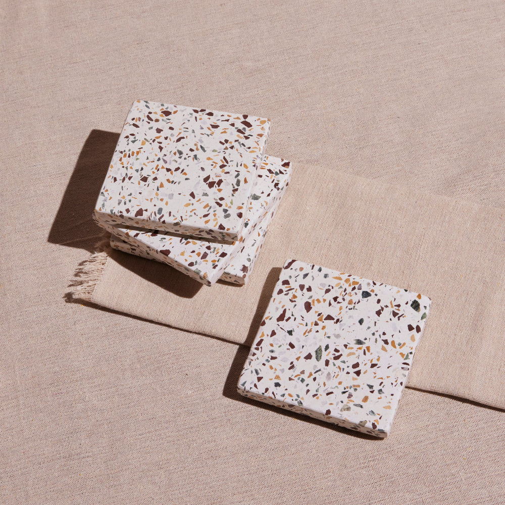 A set of four terrazzo coasters with a multicolored pattern, placed on a beige fabric surface.