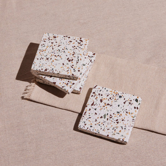 A set of four terrazzo coasters with a multicolored pattern, placed on a beige fabric surface.