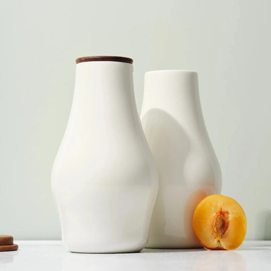 Two white ceramic carafes with a sculptural shape and a handcrafted wooden lids, one larger than the other, placed on a surface with a peach next to them.