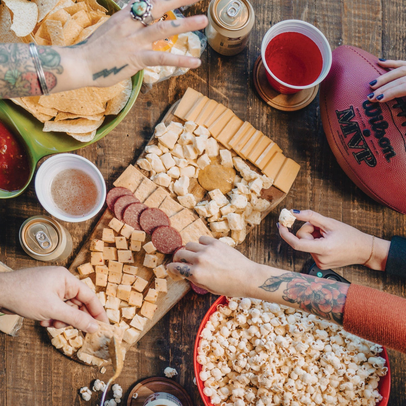 People enjoying a snack platter with chips, cheese, and drinks on a wooden table.