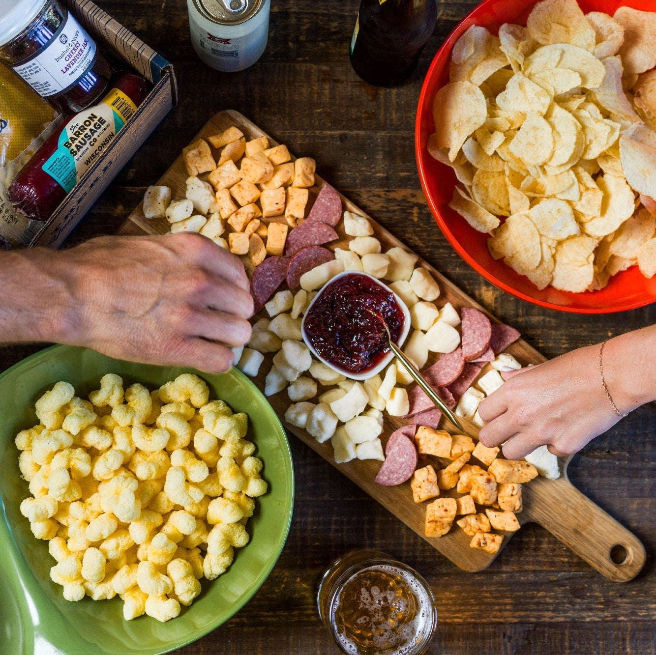 People preparing a snack platter with various food items on a wooden table.