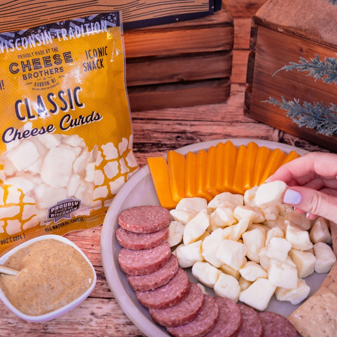 Bag of Cheese Curds with a plate of cheese curds, sliced sausages, and crackers on a wooden surface.