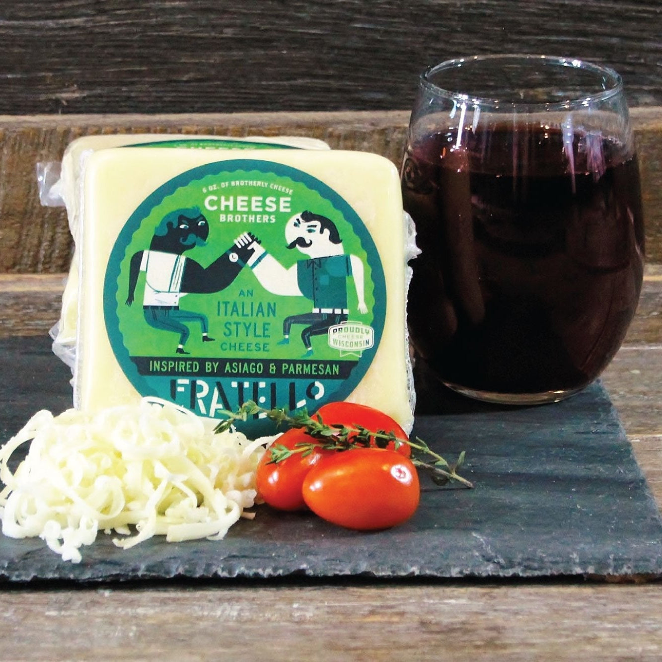 Cheese package with grated cheese, tomatoes, and a glass of dark liquid on a wooden surface.