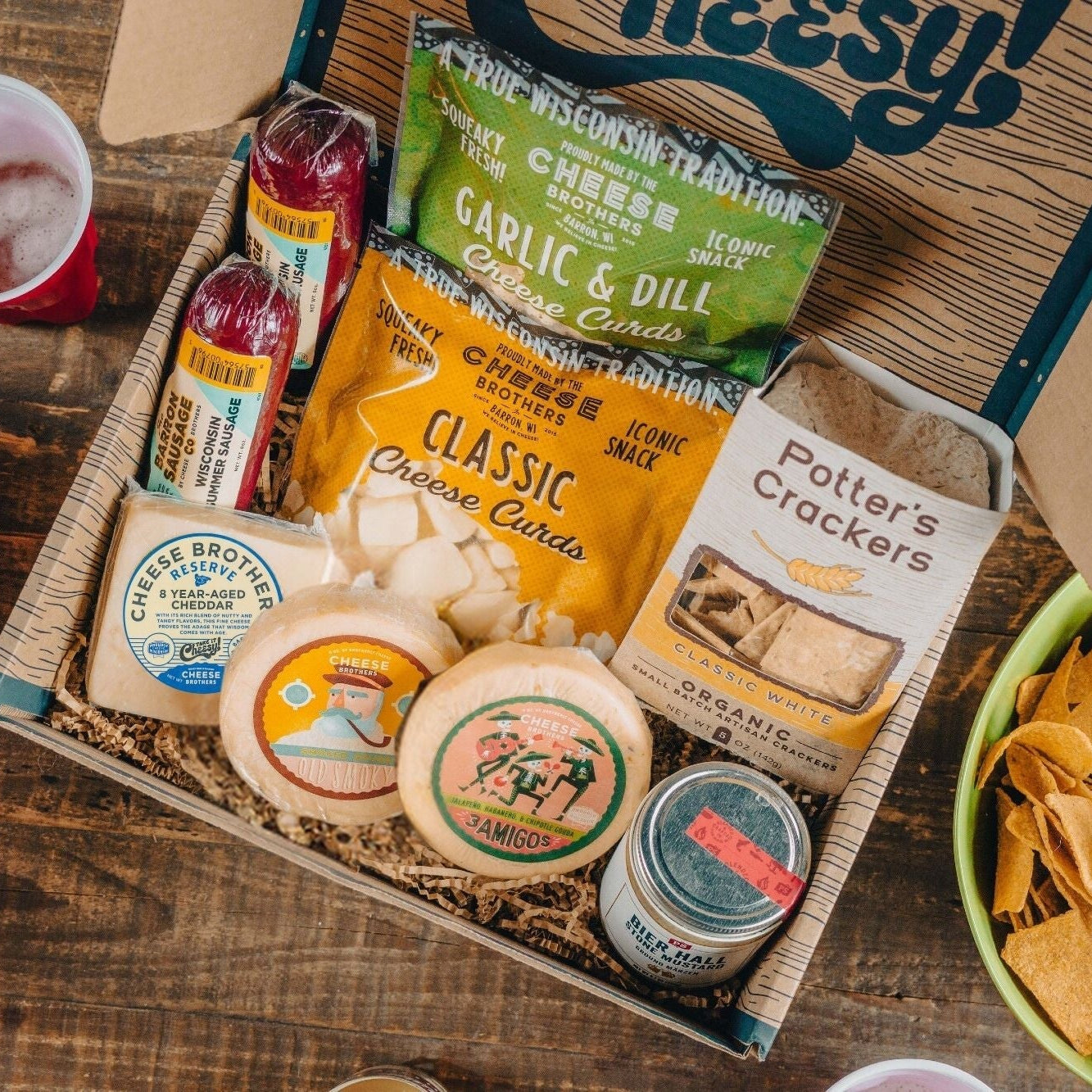 Assorted game day snacks including cheese, crackers, and chips in a box on a wooden table.