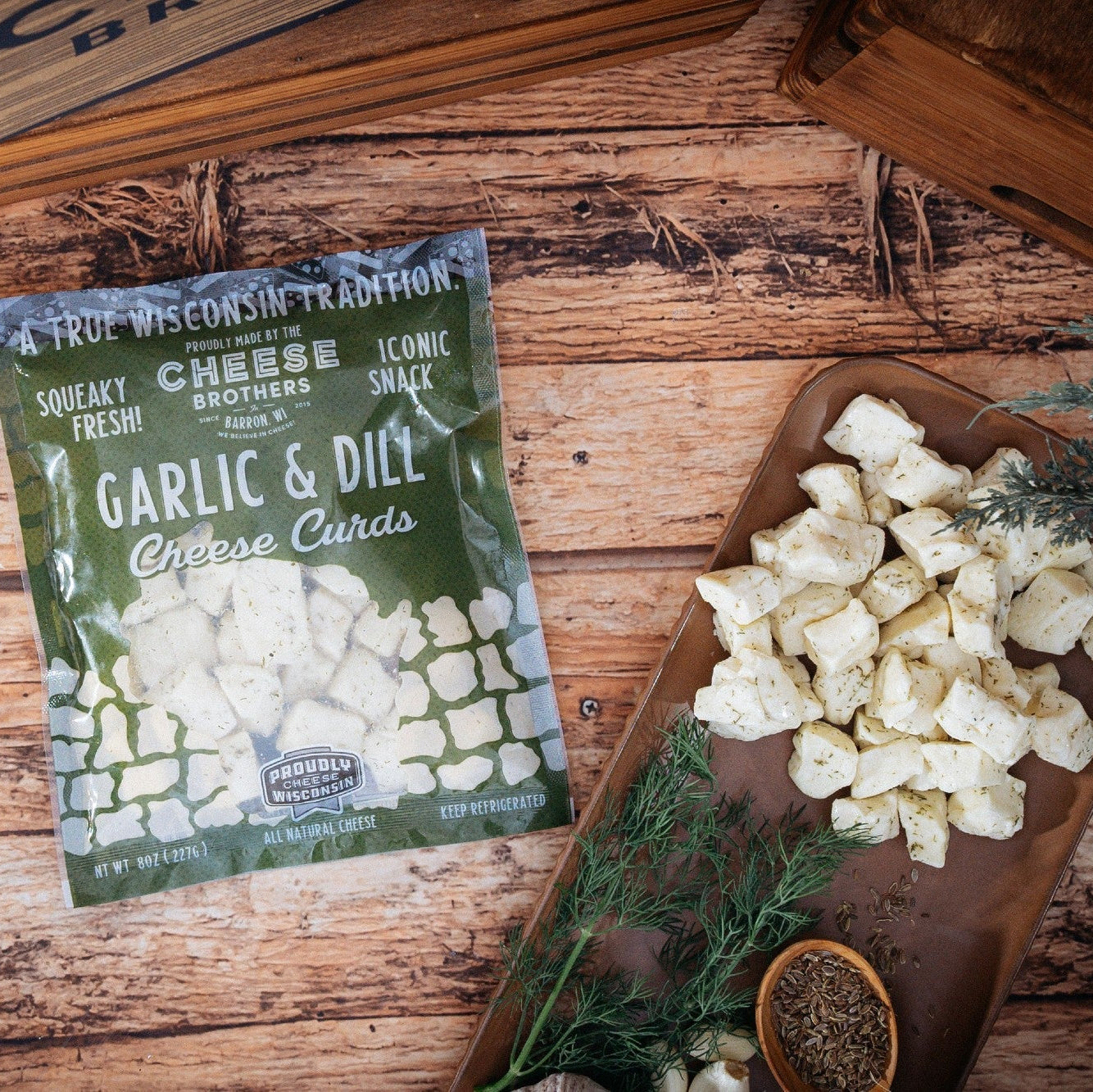 Packaged garlic and dill cheese curds on a wooden surface with herbs.