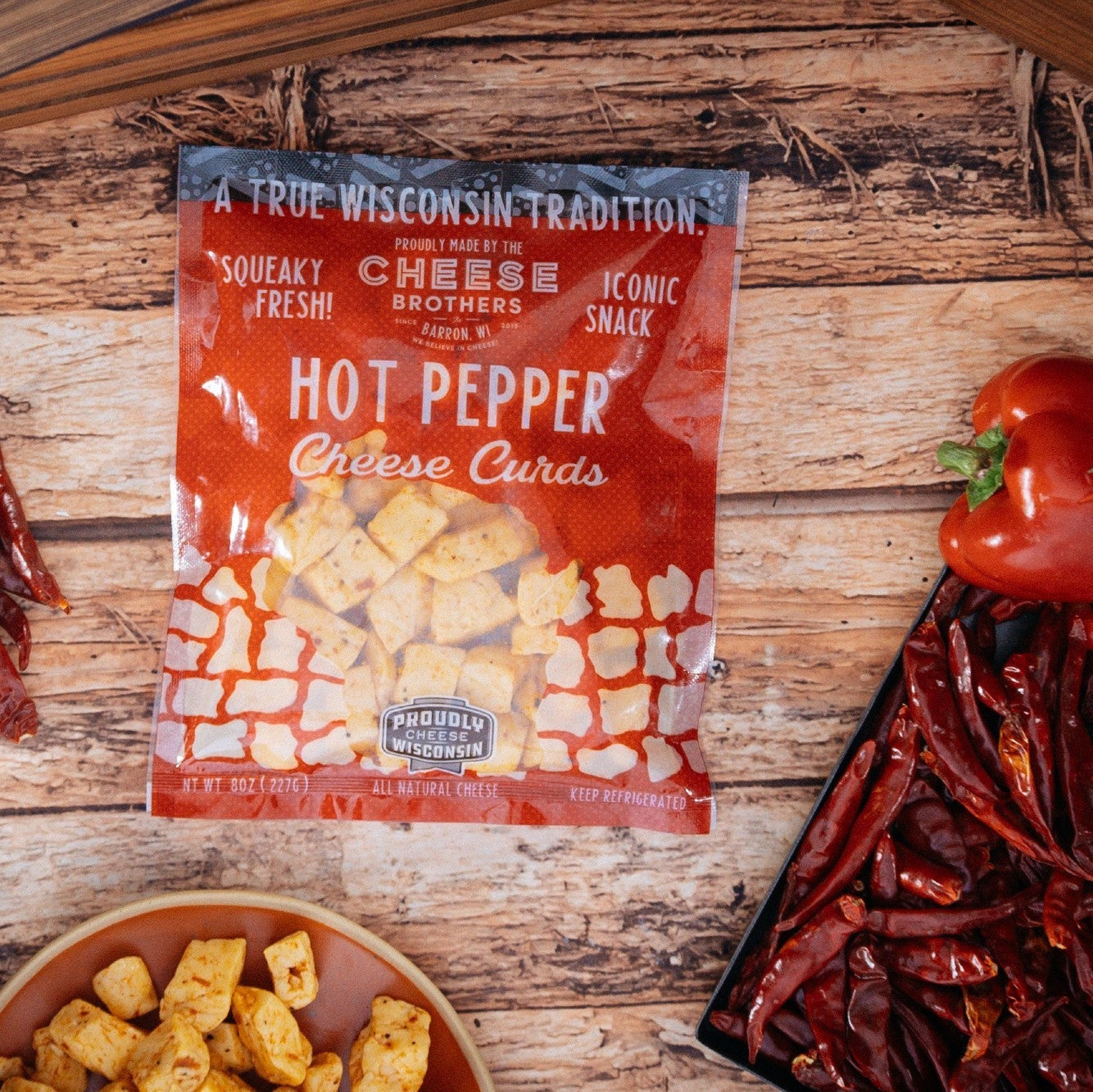 Packaged hot pepper cheese curds on a wooden surface with peppers and a bowl of cheese curds.